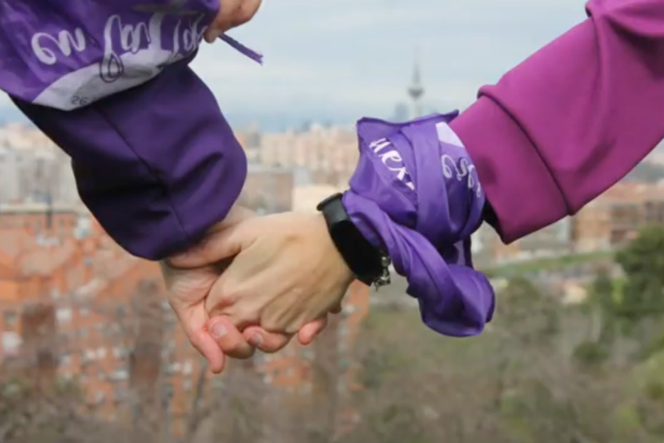Dos mujeres se dan la mano, llevando pañuelos morados de la Revuelta de Mujeres en la Iglesia